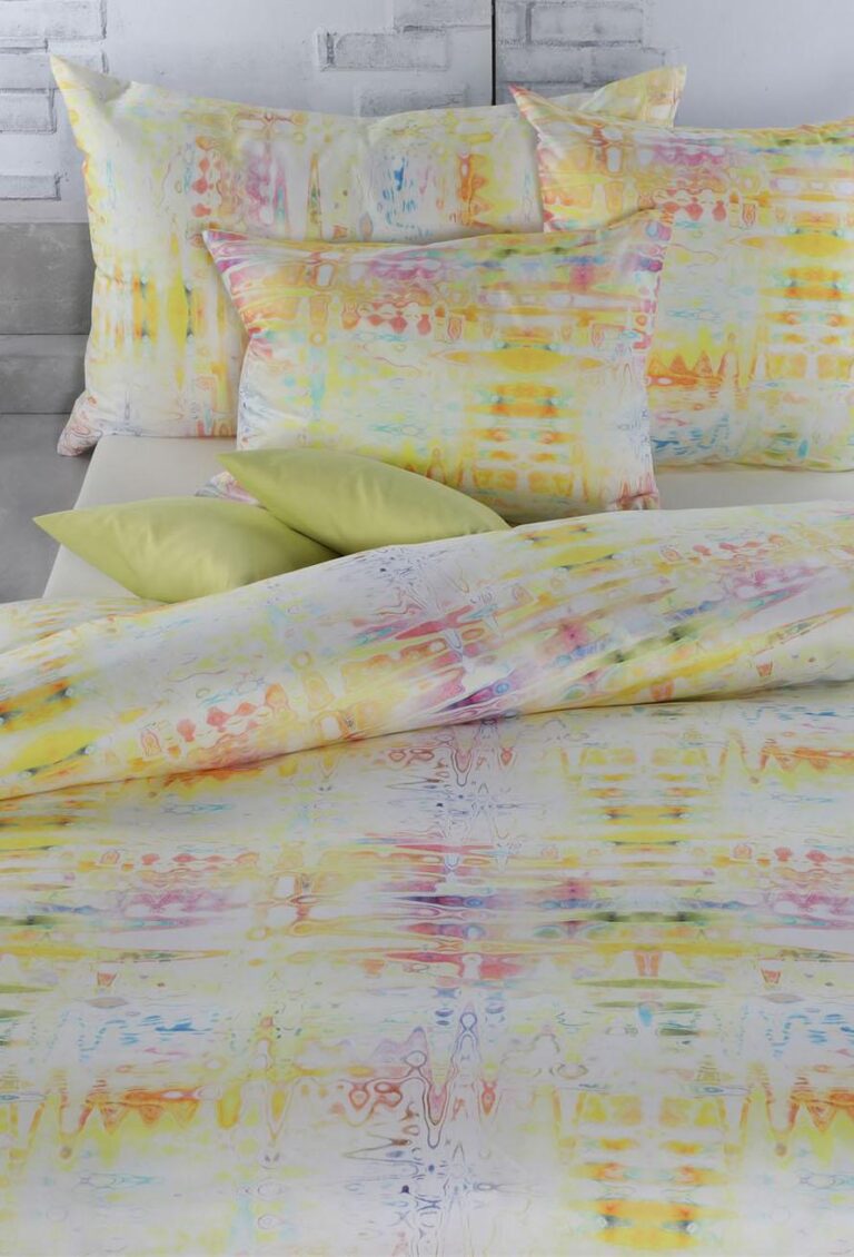 Ein Bett mit der Bettwäsche Tamara-R Selection Daisy, einer leuchtenden Bettdecke mit gelben, orangefarbenen, rosa und blauen Akzenten, steht vor einer weißen Backsteinwand und wird durch zwei hellgrüne Akzentkissen ergänzt.