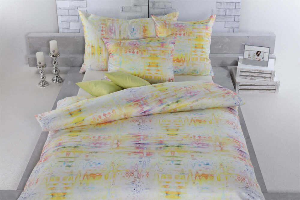 Auf einem hübsch gemachten Bett steht Bettwäsche Tamara-R Selection Daisy in pastelligen Gelb-, Orange- und Rosatönen, gepaart mit zwei grünen Akzentkissen und Accessoires wie Kerzen und einem gerahmten Foto auf einem weißen Nachttisch.