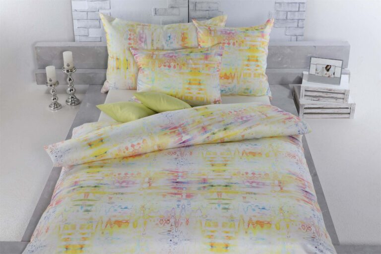 Auf einem hübsch gemachten Bett steht Bettwäsche Tamara-R Selection Daisy in pastelligen Gelb-, Orange- und Rosatönen, gepaart mit zwei grünen Akzentkissen und Accessoires wie Kerzen und einem gerahmten Foto auf einem weißen Nachttisch.