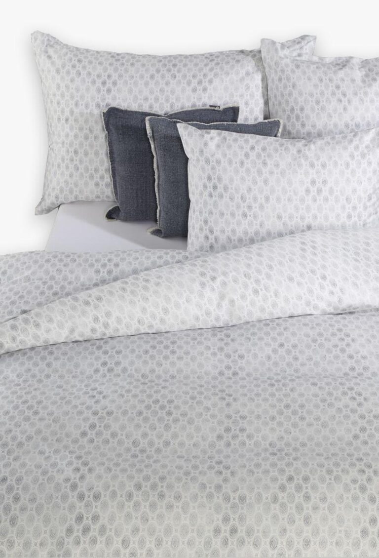 Ein ordentlich gemachtes Bett mit weißer Bettwäsche Tamara-R BeCosy Caddy blau mit dezentem Kreismuster, zwei passenden Kissen und zwei dunkelgrauen Zierkissen davor.