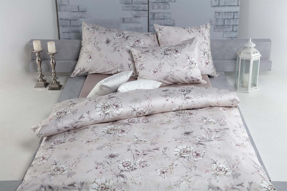 Auf einem ordentlich gemachten Bett liegt die Bettwäsche Tamara-R Selection Elissa mit hellen Blumenmustern und passenden Kissen vor einer grauen Wand mit Ziegelsteinmustern. Silberne Kerzenhalter und eine weiße Laterne sorgen für Eleganz neben dem Bett.