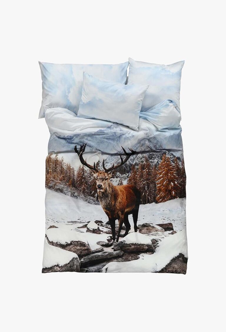 Die Bettwäsche Eugen zeigt eine lebendige Winterszene mit einem Hirsch auf verschneiten Felsen, schneebedeckten Bäumen und Bergen sowie passenden Kissen.