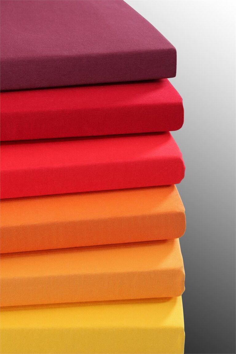 Ein Stapel von sechs rechteckigen Tamara-R Topper Fixleintuch Selection-FIX Matratzen in Farbverläufen von tiefem Violett bis zu leuchtendem Gelb, dargestellt auf einem schlichten grauen Hintergrund mit Farbverlauf.