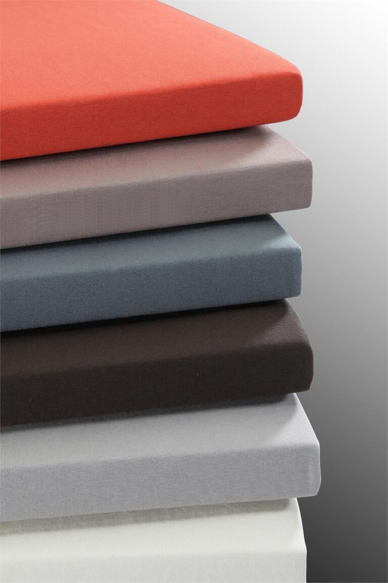 Ein Stapel von sechs Tamara-R Fixleintuch Selection-FIX Spannbetttüchern in den Farben Orange, Hellgrau, Blaugrau, Dunkelgrau, Hellgrau und Weiß ist ordentlich vor einem Hintergrund mit Farbverlauf angeordnet.