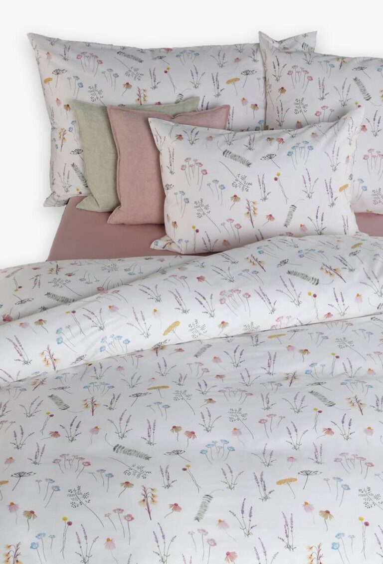 Ein ordentlich gemachtes Bett mit weißer Bettwäsche Tamara-R BeCosy Fee mit pastellfarbenen botanischen Wildblumen- und Blattdrucken, ergänzt durch mehrere koordinierte pastellfarbene Kissen, die am Kopfende des Bettes angeordnet sind.