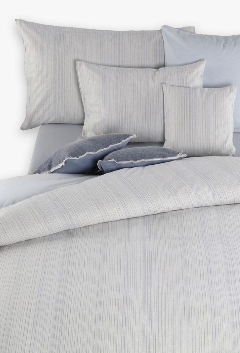 Ein ordentlich gemachtes Bett mit Bettwäsche Tamara-R BeCosy Felice in hellgrauen und blauen Nadelstreifen, komplett mit einer Bettdecke, Kissenbezügen und koordinierten einfarbigen und strukturierten Zierkissen.