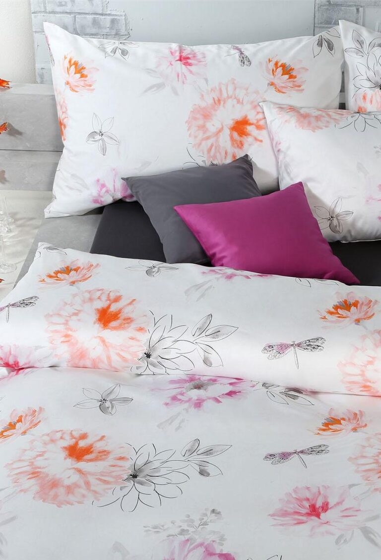Das Bett ist mit Bettwäsche Tamara-R Selection Flurina mit weißem Stoff, orangefarbenen und rosafarbenen Blumen- und Libellenmotiven ausgestattet; zwei lilafarbene Akzentkissen befinden sich zwischen passenden weißen Blumenkissen.