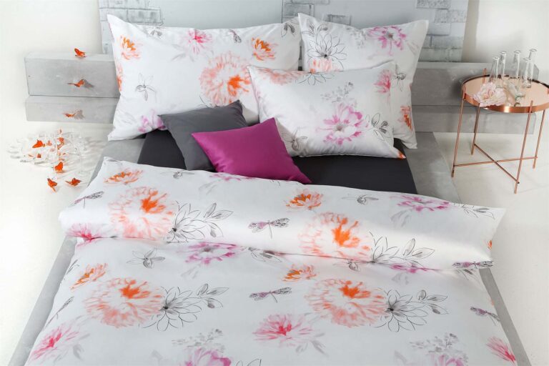 Auf einem ordentlich gemachten Bett liegt die Bettwäsche Tamara-R Selection Flurina in Weiß mit rosa und orangefarbenen Blumenmotiven. Florale, graue und magentafarbene Kissen sorgen für Charme, während auf dem Beistelltisch Gläser und orangefarbene Zierschmetterlinge stehen.
