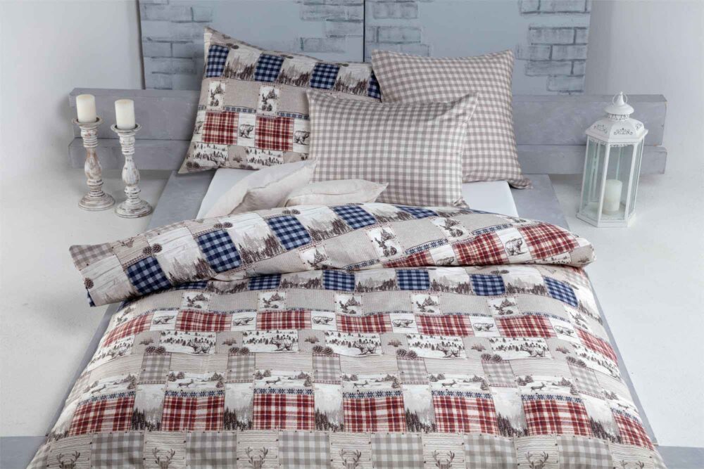 Ein hübsch gemachtes Bett mit Bettwäsche Heimatli und Kissen in Rot, Blau und Beige mit Rentier- und Karomuster. Dekorative Kerzen und eine weiße Laterne stehen neben dem Bett.