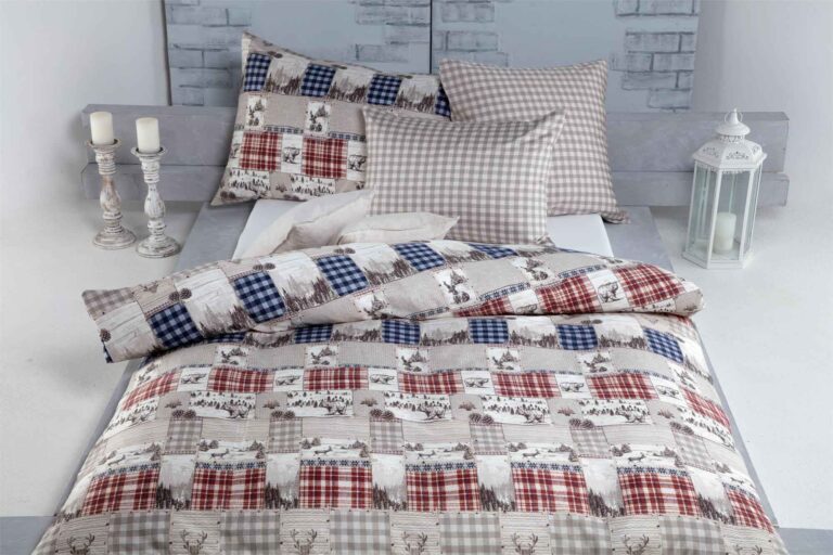 Ein hübsch gemachtes Bett mit Bettwäsche Heimatli und Kissen in Rot, Blau und Beige mit Rentier- und Karomuster. Dekorative Kerzen und eine weiße Laterne stehen neben dem Bett.