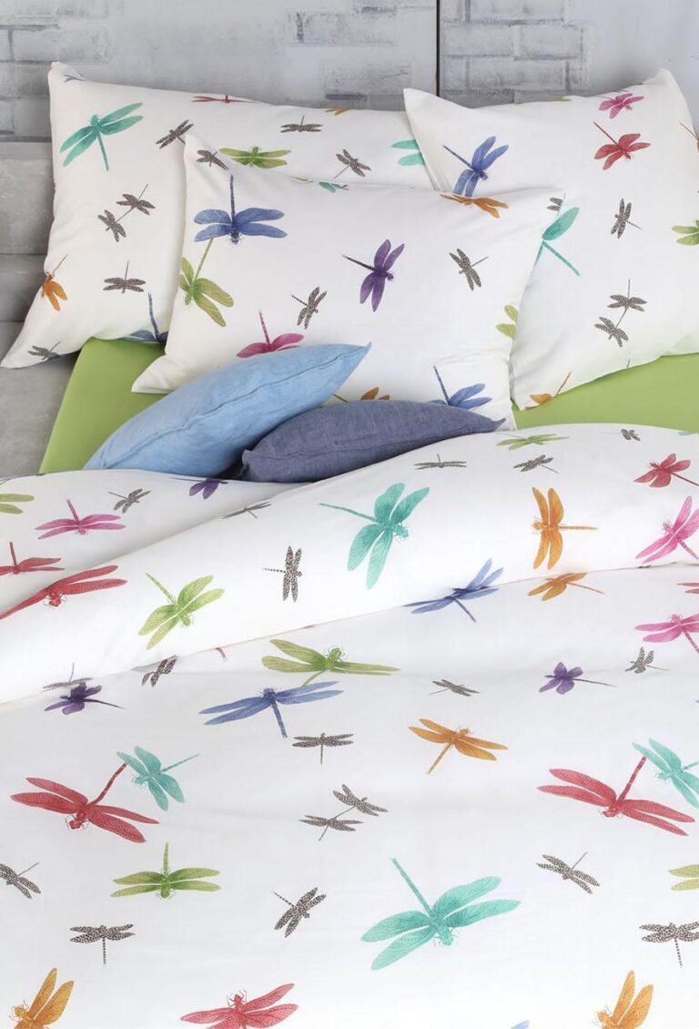 Das Bett ist mit Bettwäsche Tamara-R Selection Libella in Weiß mit bunten Libellenmotiven, blauen und grauen Kissen und einem teilweise sichtbaren grünen Bettlaken ausgestattet.