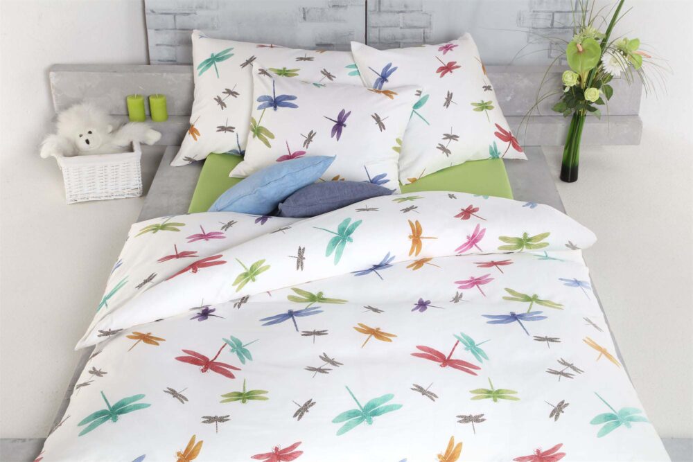Das Bett ist mit weißer Bettwäsche Tamara-R Selection Libella mit bunten Libellenmotiven bezogen; daneben stehen auf dem Nachttisch zwei grüne Tassen, ein weißer Korb mit einem Stofftier, Kissen und eine Blumenvase.