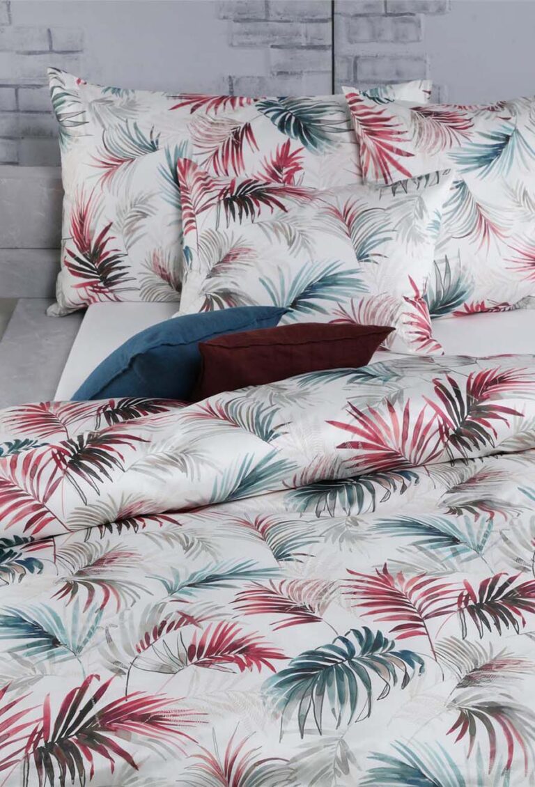Ein ordentlich gemachtes Bett mit Bettwäsche Tamara-R Selection Matteo in Weiß und einem bunten Palmenblattmuster ist mit blauen und braunen Kissen gepaart und steht vor einer hellgrauen Backsteinwand.
