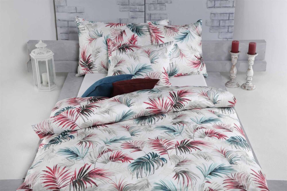 Ein Bett mit weißer Bettwäsche Tamara-R Selection Matteo mit roten, blaugrünen und grauen Palmblattmustern, zwei großen Kissen, einer Laterne auf der linken Seite und zwei roten Kerzen in weißen Haltern auf der rechten Seite vor einer hellgrauen Backsteinwand.
