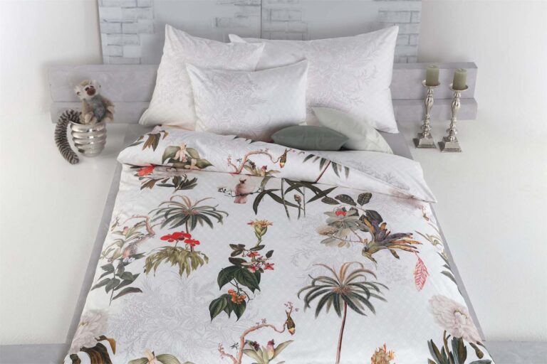 Ein ordentlich gemachtes Bett mit weißen Kissen und Bettwäsche Tamara-R Selection Paradiso, die ein lebhaftes tropisches Blumen- und Pflanzenmuster aufweist. Auf dem Nachttisch stehen zwei silberne Kerzenständer und ein Plüsch-Lemuren-Spielzeug in einer silbernen Schale.