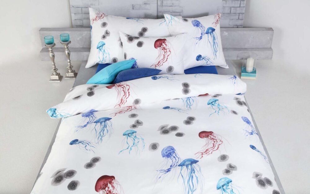 Ein ordentlich gemachtes Bett ist mit Bettwäsche Tamara-R Selection Quallina mit roten und blauen Quallen und Kreismustern bezogen. Blaue und weiße Kissen, silberne Kerzenhalter und blaue Kerzen schmücken den hellen Nachttisch.