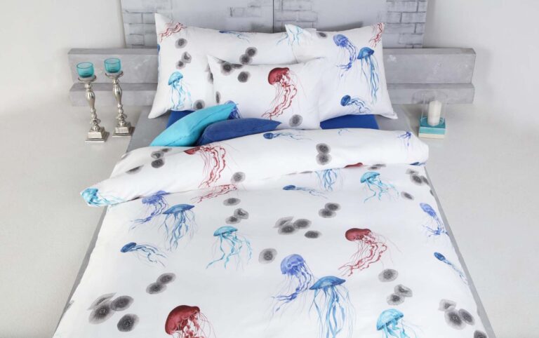 Ein ordentlich gemachtes Bett ist mit Bettwäsche Tamara-R Selection Quallina mit roten und blauen Quallen und Kreismustern bezogen. Blaue und weiße Kissen, silberne Kerzenhalter und blaue Kerzen schmücken den hellen Nachttisch.