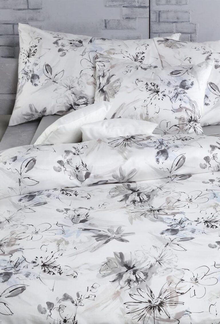 Die Bettwäsche Tamara-R Selection Ronja grau ist ein weißes Bettwäscheset mit dezenten grauen und schwarzen Blumenmustern. Der Bettbezug und die Kissen sind ordentlich auf einem Bett vor einer hellgrauen Backsteinwand angeordnet.