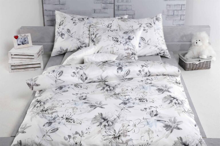 Ein ordentlich gemachtes Bett mit weißer und grauer geblümter Bettwäsche Tamara-R Selection Ronja grau, passenden Kissen und einer Steppdecke. Links stehen gestapelte weiße Kisten mit Dekoration, rechts steht ein Weidenkorb mit einem weißen Teddybär.