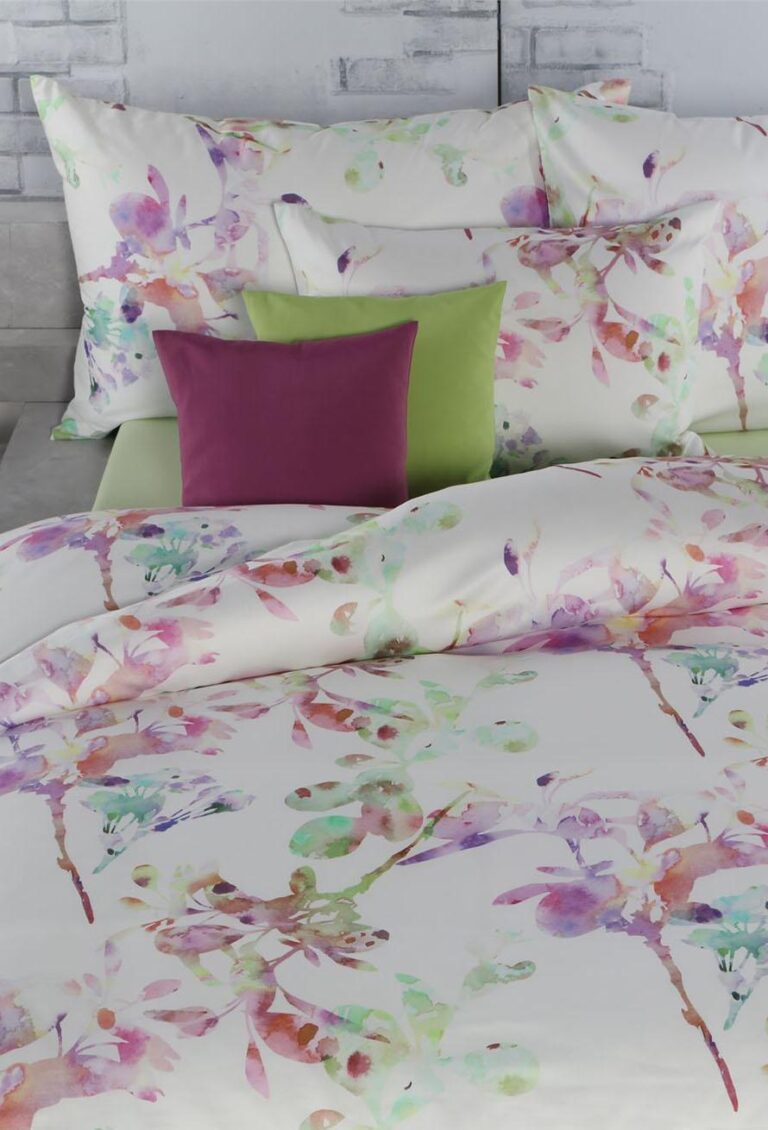 Ein Bett ist mit weißen Laken und Kissenbezügen von Bettwäsche Tamara-R Selection Silja mit einem pastellfarbenen Blumenaquarellmuster bezogen, gepaart mit grünen und pflaumenfarbenen Kissen vor einem hellgrauen Backsteinwandhintergrund.