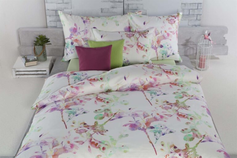 Ein Bett mit Bettwäsche Tamara-R Selection Silja - weiße Blumenbettwäsche mit pastellfarbenen Kissen - ist mit grünen und lila Kissen ausgestattet. Eine Topfpflanze und ein Vogelkäfig setzen dekorative Akzente auf jeder Seite des Bettes.