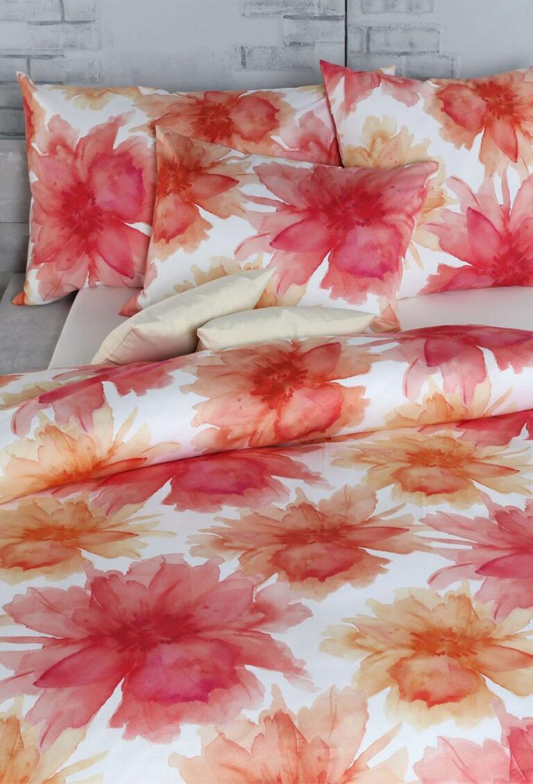 Die Bettwäsche Tamara-R Selection Sofie zeigt großflächige Blumendrucke im Aquarellstil in Rot, Orange und Gelb auf Weiß, mit passenden Kissen und Bettdecke vor einer hellen Backsteinwand.