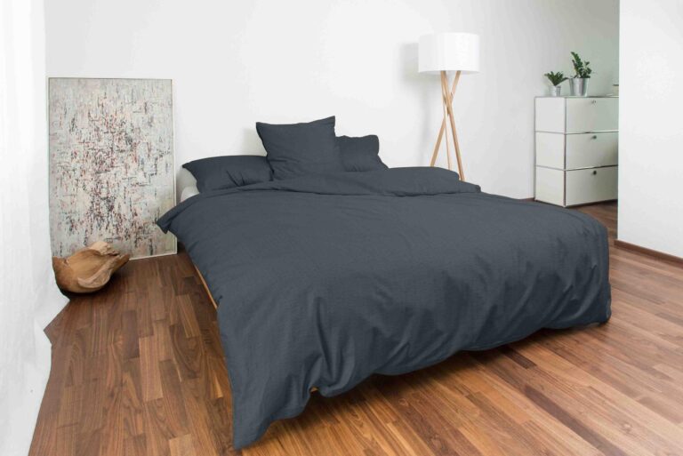 Ein minimalistisches Schlafzimmer mit einem Bett mit Bettwäsche Night Fashion Edi in Dunkelgrau, Holzböden, einer weißen Stehlampe, einer kleinen Kommode mit Pflanzen, einem abstrakten Bild an der Wand und einer hellbraunen Katze, die sich auf dem Boden zusammengerollt hat.