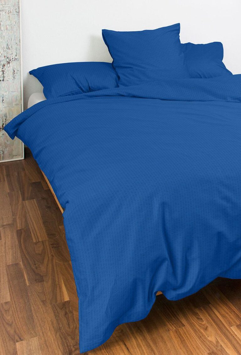 Ein Bett mit blauen Bettlaken, Kissenbezügen und einem Bettbezug von Bettwäsche Night Fashion Edi steht vor einer weißen Wand und einem Holzboden, mit vier Kissen am Kopfende und einer strukturierten Wand auf der linken Seite.