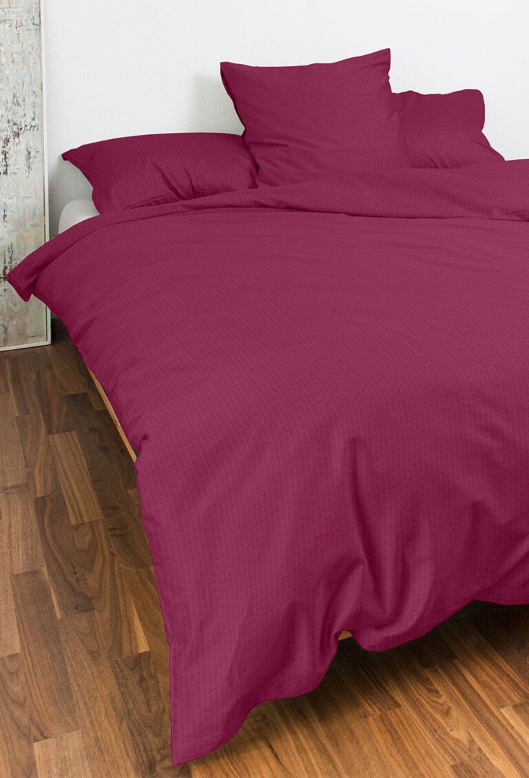 Ein stilvolles Bett in Bettwäsche Night Fashion Edi in Magenta mit passenden Kissen ruht auf einem Holzboden an einer weißen Wand, begleitet von dekorativen Kunstwerken, die in der Nähe lehnen.