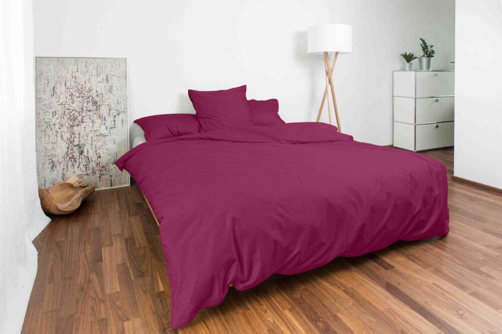 Ein ordentlich gemachtes Bett mit Bettwäsche Night Fashion Edi in Magenta steht in einem modernen Schlafzimmer mit Holzboden, weißen Wänden, einer Stehlampe, einer weißen Kommode mit Pflanzen und abstrakter Kunst an der Wand.