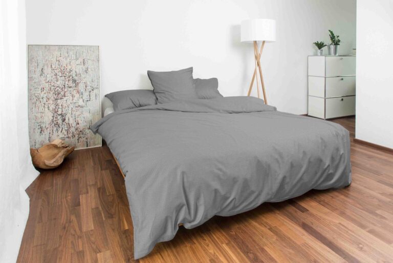 Ein modernes Schlafzimmer verfügt über ein ordentlich gemachtes Bett mit Bettwäsche Night Fashion Edi, Holzböden, ein abstraktes Gemälde an der Wand, eine Stehlampe, eine Kommode und Topfpflanzen in der Ecke.