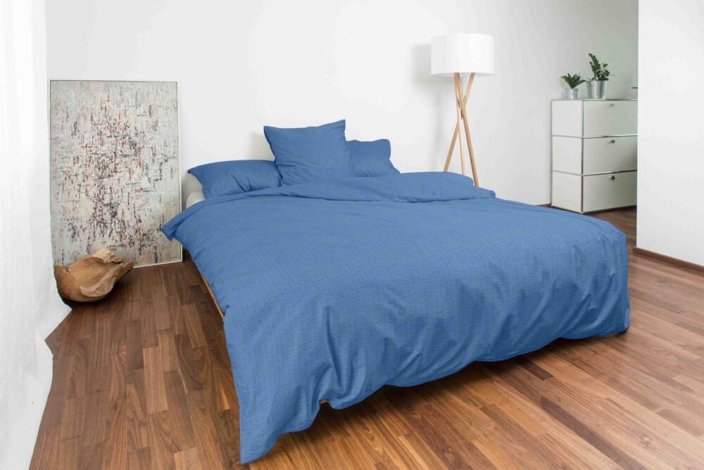 In einem minimalistischen Schlafzimmer mit Holzfußboden stehen ein ordentlich gemachtes Bett in Bettwäsche Night Fashion Edi, eine Stehlampe, Topfpflanzen auf einer weißen Kommode, abstrakte Kunst an der Wand und eine hellbraune Katze, die sich in der Nähe zusammengerollt hat.