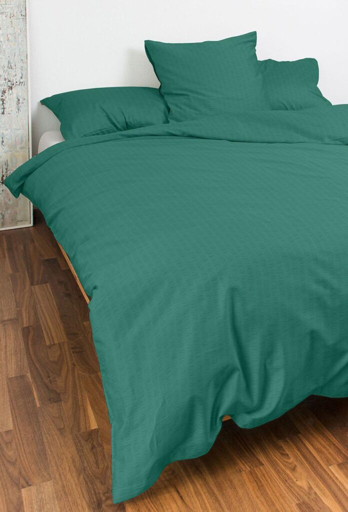 Ein ordentlich gemachtes Bett mit Bettwäsche Night Fashion Edi in Teal und passenden Kissen steht vor einer weißen Wand und einem Holzboden. Die Bettdecke ist über die Bettseite drapiert, und im Hintergrund ist teilweise abstrakte Wandkunst zu sehen.