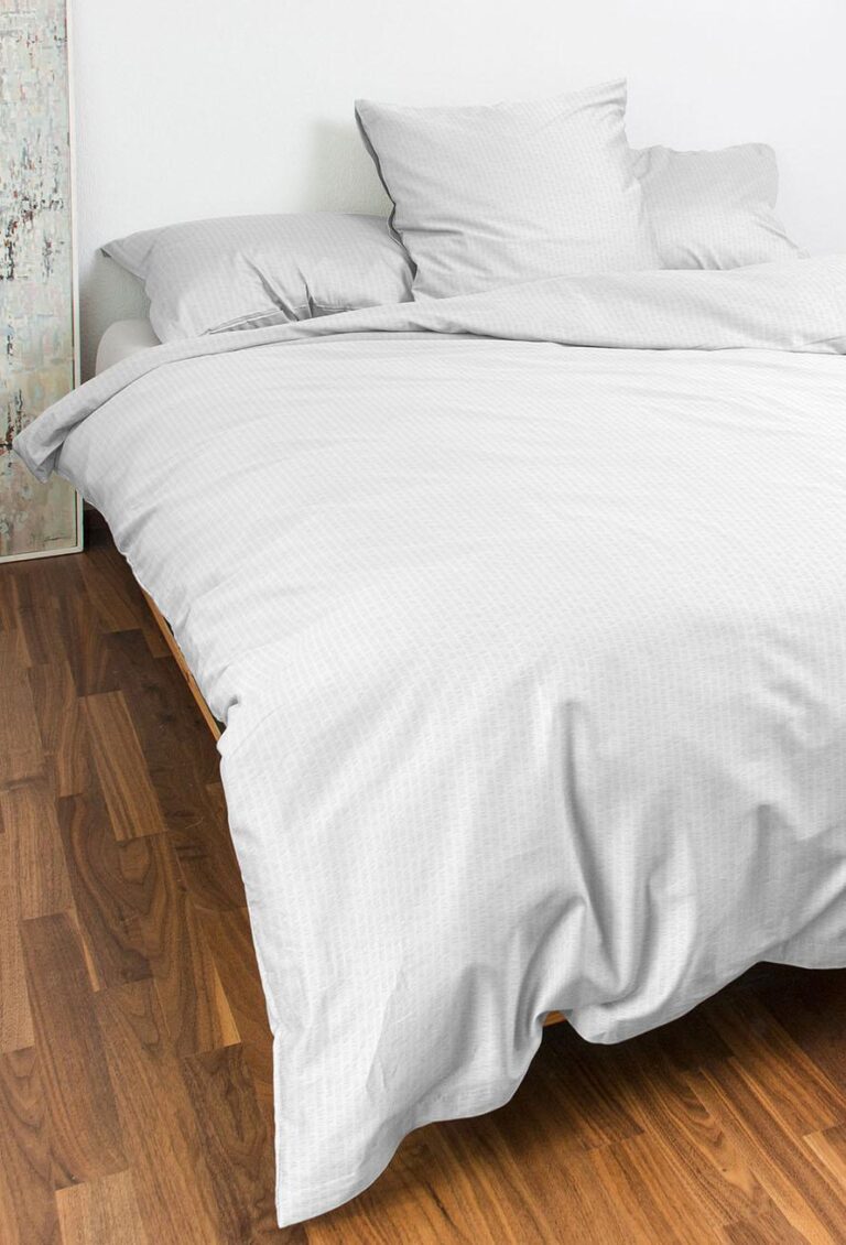 Ein ordentlich gemachtes Bett mit Bettwäsche Night Fashion Edi in Weiß und Hellgrau, mit passenden Kissen und Bettdecke, steht auf einem Holzboden neben einer weißen Wand und einer dekorativen Tafel.