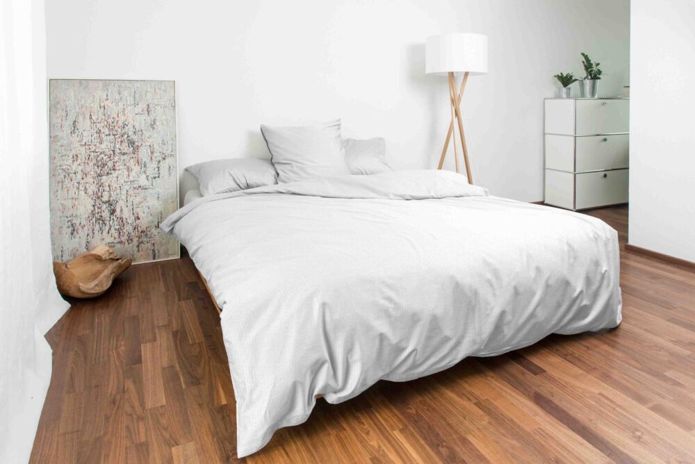 Ein minimalistisches Schlafzimmer mit einem niedrigen Bett mit Bettwäsche Night Fashion Edi, einer modernen Stehlampe, einer weißen Kommode mit Pflanzen, Holzboden und einem abstrakten Gemälde, das an der Wand lehnt.