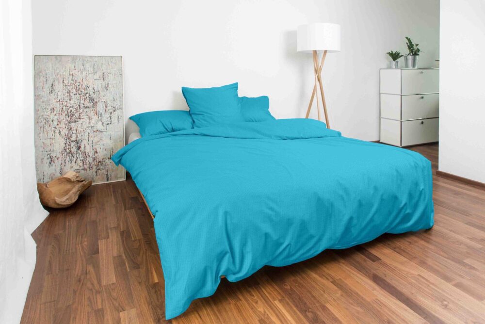 Ein modernes Schlafzimmer mit Bettwäsche Night Fashion Edi auf einer türkisfarbenen Tagesdecke, zwei Kissen, Holzboden, weißen Wänden, einer Stehlampe, einer Kommode mit Pflanzen, abstrakter Kunst und einer Katze, die sich an der Wand zusammengerollt hat.