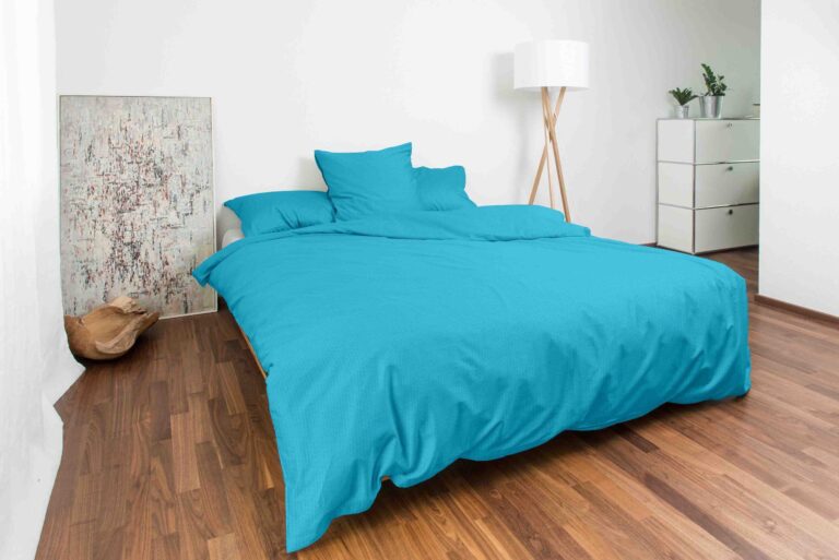 Ein modernes Schlafzimmer mit Bettwäsche Night Fashion Edi auf einer türkisfarbenen Tagesdecke, zwei Kissen, Holzboden, weißen Wänden, einer Stehlampe, einer Kommode mit Pflanzen, abstrakter Kunst und einer Katze, die sich an der Wand zusammengerollt hat.