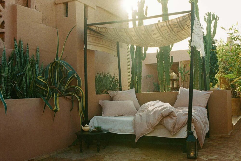 Ein gemütliches Outdoor-Himmelbett mit Bettwäsche Journey Living Bellecour ruht auf einer gefliesten Terrasse zwischen hohen Kakteen und Grünpflanzen, neben rosafarbenen Stuckwänden im warmen Sonnenlicht. Eine Laterne und Topfpflanzen vervollständigen die einladende Szene.