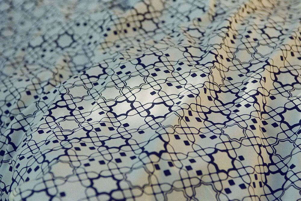 Nahaufnahme eines Stoffes mit einem komplizierten, sich wiederholenden geometrischen Muster in dunklen Linien auf hellem Grund, das weiche Falten und Knicke zeigt.