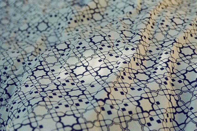 Nahaufnahme eines Stoffes mit einem komplizierten, sich wiederholenden geometrischen Muster in dunklen Linien auf hellem Grund, das weiche Falten und Knicke zeigt.