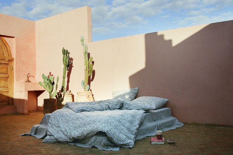 Ein gemütliches Bett mit gemustertem Bettzeug steht im Freien an einer rosafarbenen Wand, neben zwei getopften Kakteen, unter einem blauen Himmel mit vereinzelten Wolken. Ein Tablett mit einer Tasse und einem Buch steht neben dem Bett auf dem gefliesten Boden.