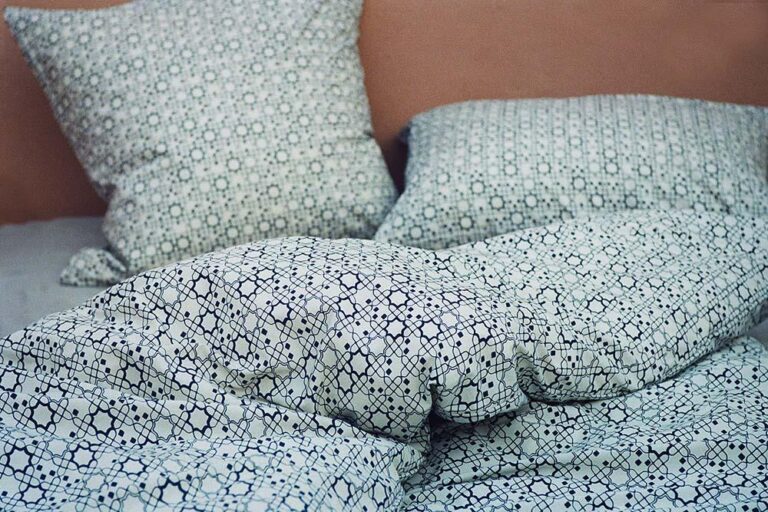 Ein Bett mit zwei Kissen und einer Steppdecke, alle mit weißem Stoff mit einem sich wiederholenden schwarzen geometrischen Muster bezogen. Die Kissen und die Bettdecke wirken leicht zerknittert, was auf eine kürzliche Benutzung schließen lässt.