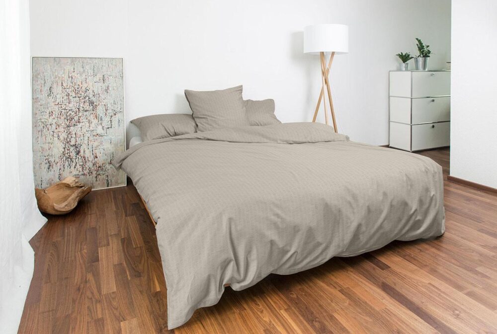 Ein modernes Schlafzimmer mit einem sauber gemachten Bett aus Bettwäsche Night Fashion Edi, Holzböden, abstrakten Kunstwerken an den Wänden, einer hohen Stehlampe, einer weißen Kommode und Topfpflanzen in der Ecke.