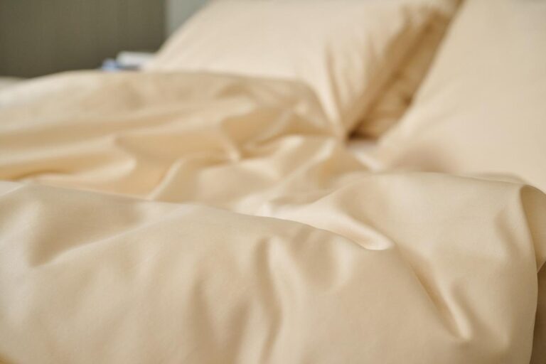 Nahaufnahme eines Bettes mit Bettwäsche Journey Living Braga Sand, mit hellbeigen oder cremefarbenen Laken und Kissen aus weichem, leicht geknittertem Stoff.