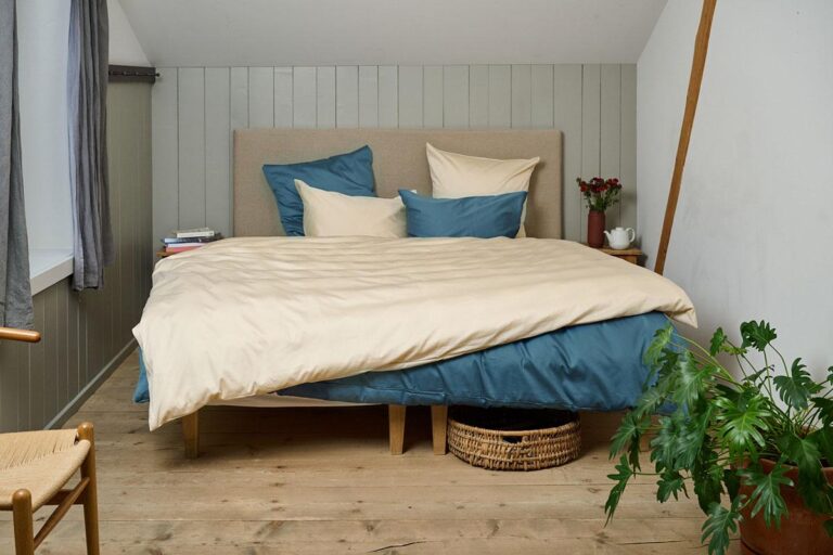 Ein gemütliches Schlafzimmer mit einem großen Bett in Bettwäsche Journey Living Braga Sand, Holzfußboden, einer Topfpflanze im Vordergrund, einem Weidenkorb unter dem Bett und einem kleinen Tisch links mit Büchern, Blumen und einer Tasse.