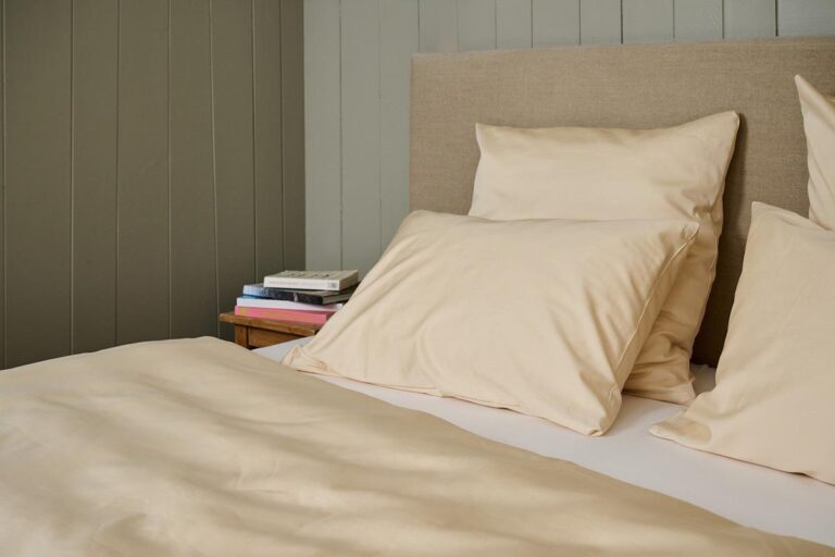 Ein ordentlich gemachtes Bett mit Bettwäsche Journey Living Braga Sand und beigen Kissen steht neben einem hölzernen Nachttisch mit Büchern, alles vor einer grau getäfelten Wand.