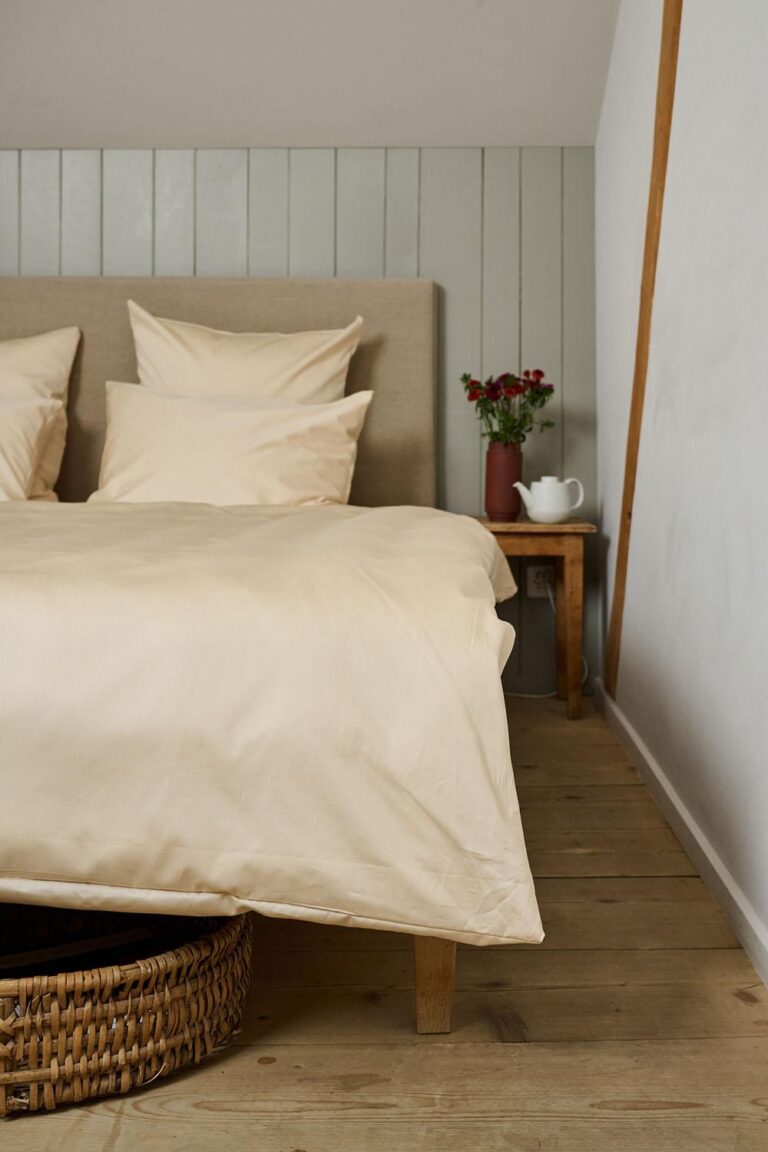 In einem gemütlichen Zimmer steht ein ordentlich gemachtes Bett mit der Bettwäsche Journey Living Braga Sand und mehreren Kissen. Auf einem hölzernen Nachttisch stehen eine Vase mit roten Blumen sowie eine weiße Teekanne und Tasse. Boden und Wand sind mit hellem Holz verkleidet.