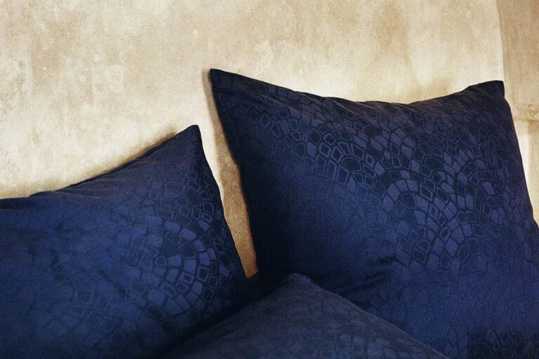 Drei dunkelblaue Bettwäsche Journey Living Casablanca-Kissen mit einem subtilen geometrischen Muster sitzen vor einer strukturierten beigen Wand, zwei überlappend und eines leicht zurückliegend angeordnet.