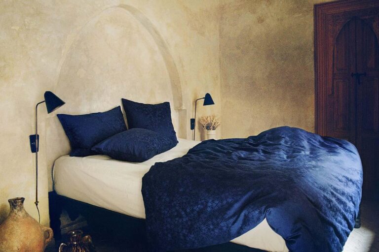 Ein gemütliches Schlafzimmer mit einer strukturierten beigen Wand, einem weißen Bett mit der dunkelblauen Bettwäsche Journey Living Casablanca und Kissen, zwei schwarzen Wandlampen und rustikalem Dekor, darunter ein großer Keramikkrug auf dem Boden.