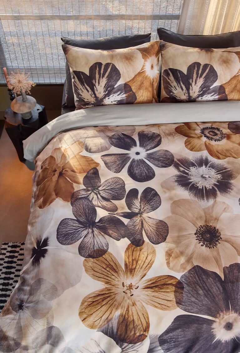 Das Bett ist mit Bettwäsche von Beddinghouse Jane mit einem auffälligen Blumendruck in Braun, Beige und Schwarz ausgestattet. Das Zimmer hat einen Nachttisch mit Dekoration und Sonnenlicht, das durch die Jalousien fällt.